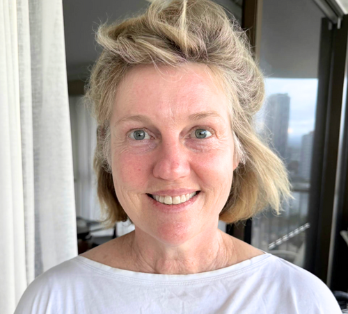 Woman with blonde hair styled in an updo, wearing a white top, standing indoors.