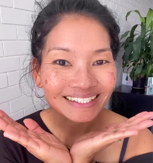 Woman with a smile, showing her hands in front of her face, with a plant in the background.